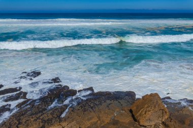 Güçlü dalgalar, dalgalı mavi gökyüzünün altında engebeli kıyı kayalarına çarpar. Okyanusa bakan uzak bir uçurum binası vardır.