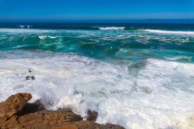 Güçlü dalgalar, dalgalı mavi gökyüzünün altında engebeli kıyı kayalarına çarpar. Okyanusa bakan uzak bir uçurum binası vardır.