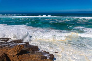 Güçlü dalgalar, dalgalı mavi gökyüzünün altında engebeli kıyı kayalarına çarpar. Okyanusa bakan uzak bir uçurum binası vardır.