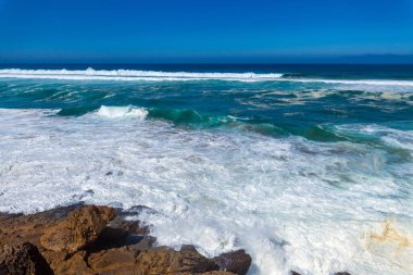 Güçlü dalgalar, dalgalı mavi gökyüzünün altında engebeli kıyı kayalarına çarpar. Okyanusa bakan uzak bir uçurum binası vardır.