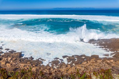 Güçlü dalgalar, dalgalı mavi gökyüzünün altında engebeli kıyı kayalarına çarpar. Okyanusa bakan uzak bir uçurum binası vardır.