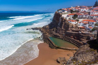 A vibrant coastal village perched on cliffs overlooks a sandy beach, turquoise waves, and a natural rock pool under a clear blue sky in  Miradouro das Azenhas do Mar