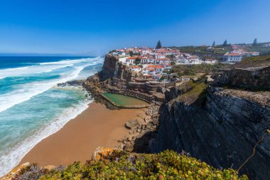 A vibrant coastal village perched on cliffs overlooks a sandy beach, turquoise waves, and a natural rock pool under a clear blue sky in  Miradouro das Azenhas do Mar