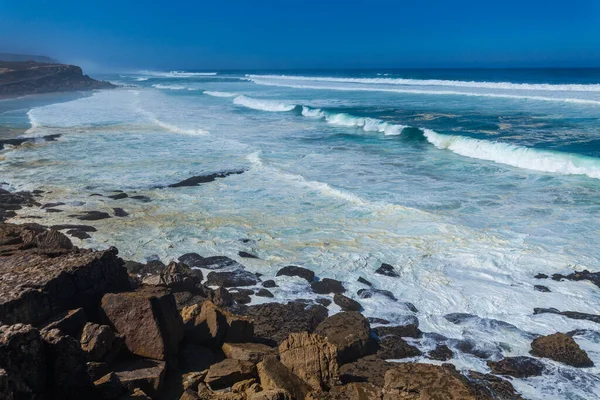 Turkuaz dalgaların kayalık bir sahile çarptığı çarpıcı bir sahil sahnesi, Apple sahilinde canlı mavi bir gökyüzünün altında kumlu kumsalla buluşması.