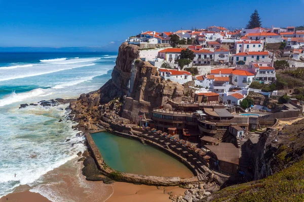 A vibrant coastal village perched on cliffs overlooks a sandy beach, turquoise waves, and a natural rock pool under a clear blue sky in  Miradouro das Azenhas do Mar