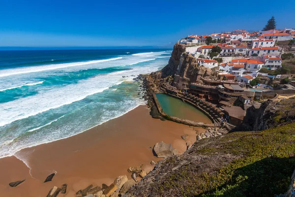 A vibrant coastal village perched on cliffs overlooks a sandy beach, turquoise waves, and a natural rock pool under a clear blue sky in  Miradouro das Azenhas do Mar