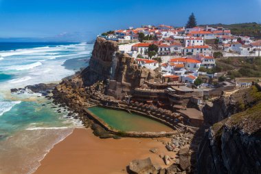 A vibrant coastal village perched on cliffs overlooks a sandy beach, turquoise waves, and a natural rock pool under a clear blue sky in  Miradouro das Azenhas do Mar