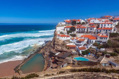 A vibrant coastal village perched on cliffs overlooks a sandy beach, turquoise waves, and a natural rock pool under a clear blue sky in  Miradouro das Azenhas do Mar