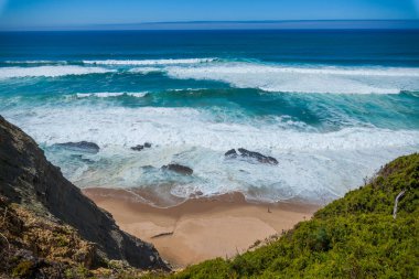 A stunning coastal view from above, showcasing turquoise waves crashing onto a sandy beach nestled between rocky cliffs and green shrubs under a clear blue sky in v