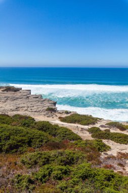 Praia da Aguda 'da açık mavi bir gökyüzü altında yemyeşil çalılar ve kumlu arazilerle çevrili kayalık kayalıklara çarpan turkuaz okyanus dalgalarını gösteren nefes kesici bir kıyı manzarası.
