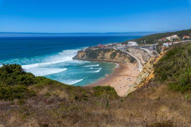 A stunning coastal panorama with turquoise waves crashing onto a sandy beach, framed by cliffs, greenery, and scattered buildings under a vibrant blue sky in Praia do Magoito