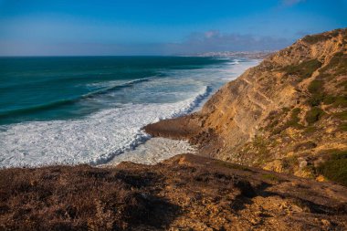 Sessiz bir kıyı yolu yemyeşil bitki örtüsü ve uzun kamışların arasından Atlantik Okyanusu 'ndaki açık mavi gökyüzünün altına doğru uzanır.