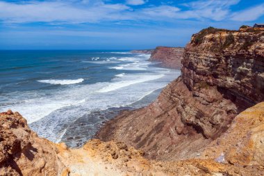 Parlak mavi gökyüzü altında yükselen kayalıklara çarpan güçlü dalgalarla çarpan dramatik bir kıyı manzarası, Praia da Escadinha 'da engebeli araziler ve doğal aşınmalar sergiliyor.