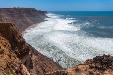 Güçlü dalgaların kumlu bir sahile çarptığı nefes kesici bir kıyı manzarası, yüksek uçurumlar ve Praia da Escadinha 'da açık mavi bir gökyüzü tarafından çerçevelenmiş.