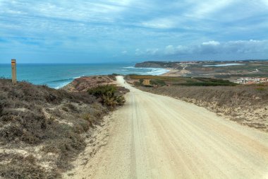 Praia Azul 'da kıyı boyunca uzanan dolambaçlı kumlu bir yol kıyıya vuran dalgalarla okyanusa doğru ilerliyor. Kuru bitki örtüsü ve bulutlu bir gökyüzünün altındaki uzak binalarla çevrili.