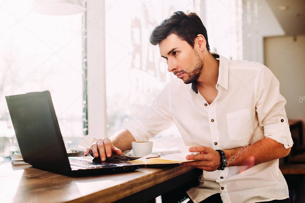 Young male freelancer chatting on mobile phone while sitting front open laptop computer in natural food cafe, handsome man reading text message during work on net-book in comfortable coffee shop