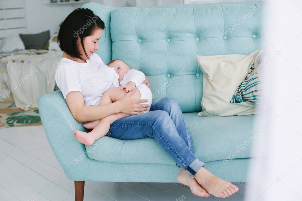 Young mother sits on the sofa in the bedroom and breastfeeds her one year old baby