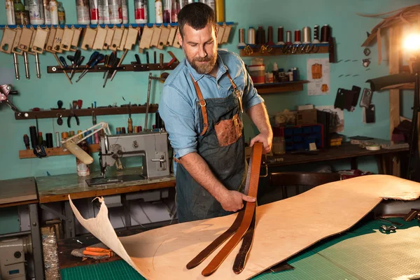 Artesano en delantal trabajando con cuero en el taller, proceso de fabricación de la correa . — Foto de Stock