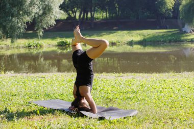 Genç ve çekici bir kadın yoga yapıyor, amuda kalkma egzersizi yapıyor, sirsasana pozisyonunda baddha konasana yapıyor, spor yapıyor, siyah spor kıyafetleri giyiyor, parkta. Arka plan