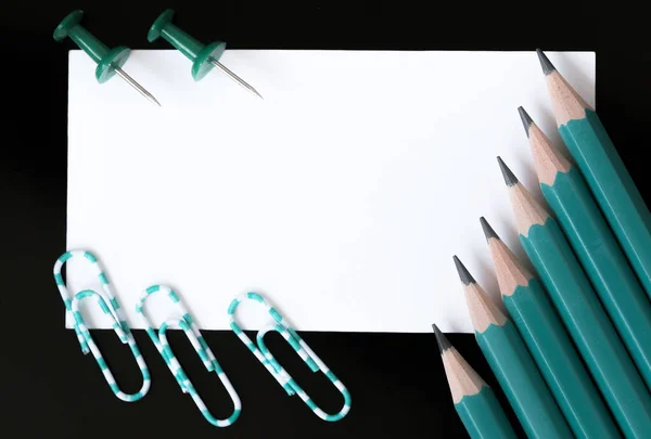 White rectangular sheet of note paper with pencils and paper clips close-up on a black background macro photography