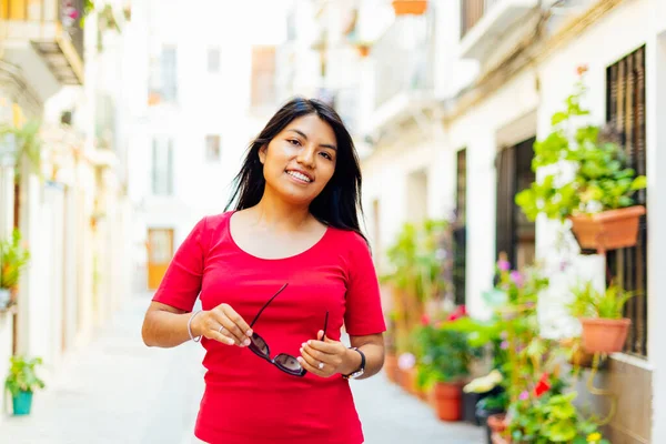 Young latin brunette girl looking at camera with sunglasses in hands on bucolic street in European city. urban tourism concept