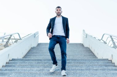 Ceketli ve kot pantolonlu sıradan bir genç adam merdivenlerden iniyor, açık havada.