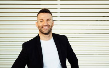 Siyah ceketli ve kısa sakallı beyaz tişörtlü genç adam çizgili arka planda kameraya gülümsüyor.