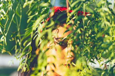 Siyah saçlı, kırmızı kurdeleli, kafası ağaç yapraklarının arasında olan ve kameraya bakan genç Latin kızın yakın çekimi.