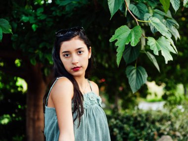 Uzun siyah saçlı, mavi elbiseli, kafasında güneş gözlüğü olan, bitkilerin arasında kameraya bakan Latin bir genç kız. Boşluğu kopyala