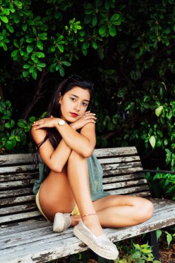 Uzun siyah saçlı ve mavi elbiseli Latin genç kız, çapraz kollu, ahşap bankta oturuyor yeşillikler arasında, kameraya bakıyor, romantik konsept