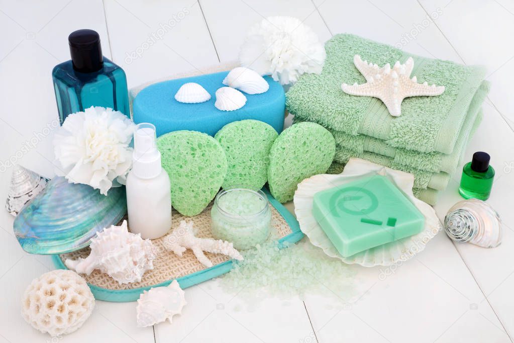 Spa and bathroom beauty treatment accessories with tea tree soaps, body lotion, blue bath foam, aromatherapy essential oil, sponges, wash cloths, ex foliating scrub and bath salts with decorative seashells on white wood background.