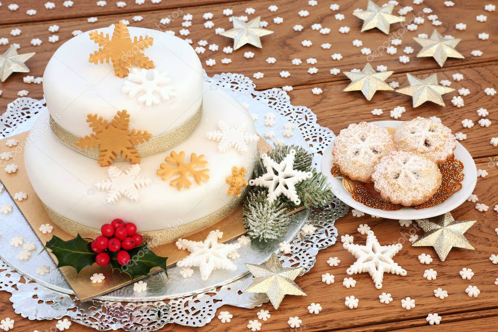 Iced Christmas cake with mince pies, snowflake decorations, gold stars, winter holly and fir on rustic oak background.