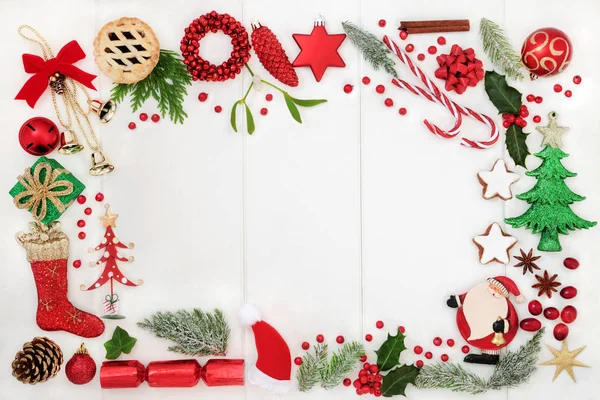 Christmas background border with traditional symbols of bauble decorations, candy canes, mince pies, fruit, spices and winter flora on rustic white wood. Top view.