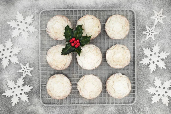 Freshly baked Christmas mince pies on a baking rack with icing sugar dusting and snowflake bauble decorations with holly berry leaf sprig. Top view.