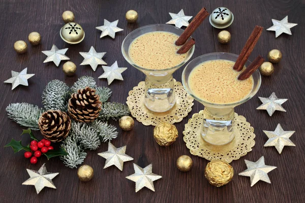 Traditional eggnog Christmas drink with gold star and bell decorations, foil wrapped chocolate balls, with winter flora of holly, fir and pine cones on rustic oak table background.