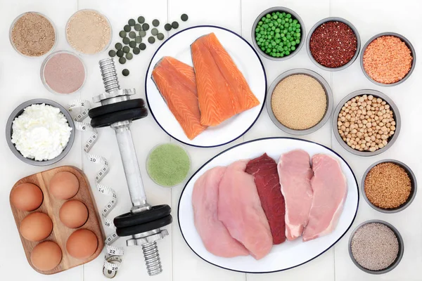 Health food for body builders high in protein including dietary supplement powders, fish, meat, dairy, pulses, grains and seeds with weights and tape measure on white wood background.