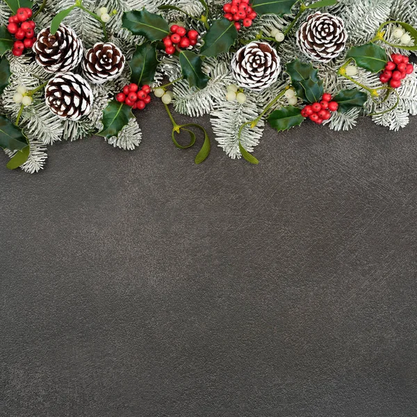 Traditional natural winter solstice snow covered spruce fir border with holly, mistletoe & pine cones on grey grunge background. Festive design for Christmas & New Year holiday season. Flat lay, copy space.
