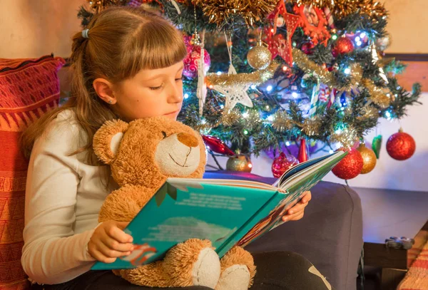 若い女の子は彼女のテディベアとクリスマス ツリーの前で本を読みます — ストック写真