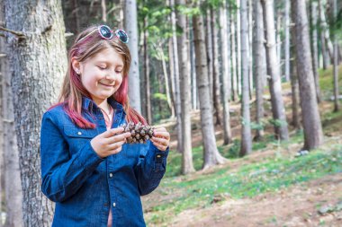 ormanda bir çam konisi tutan gülümseyen küçük kız