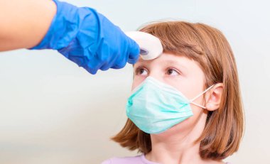 Çocuk doktoru ya da doktor, ilkokul çağındaki kızın vücut ısısını virüs belirtileri için kızılötesi alın termometresi (termometre tabancası) kullanarak kontrol eder - salgın koronavirüs salgını kavramı