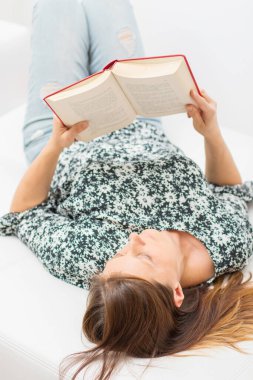 Güzel bir kız beyaz bir kanepede kitap okuyor.