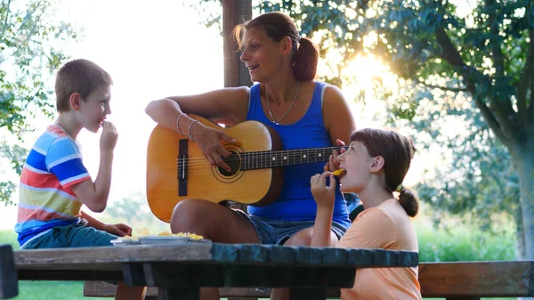 Mom and her two children sing songs together playing guitar outdoors on a sunny day. Concept of relaxation and healthy life