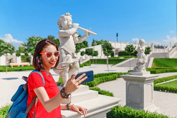 年轻女性旅行者制作自拍与智能手机与丘比特雕像在夏季花园或公园 — 图库照片