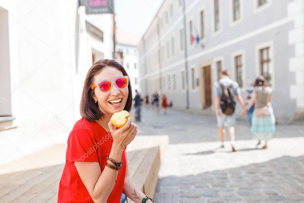 Attractive young tourist woman having snack and resting on the street and eating an apple