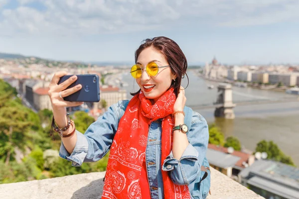 快乐的年轻亚洲妇女旅游制作自拍照片使用她的智能手机站在布达佩斯城市的旧桥前与议会大厦的看法 — 图库照片