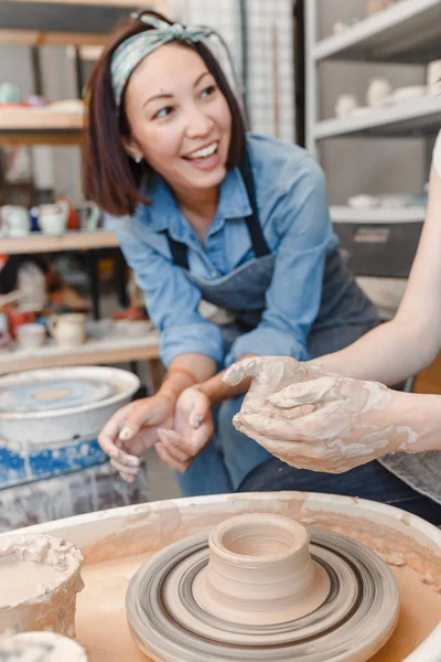 Two girls friends smiling and talking while working on potters wheel making clay handmade craft in pottery workshop, friendship and guidance concept