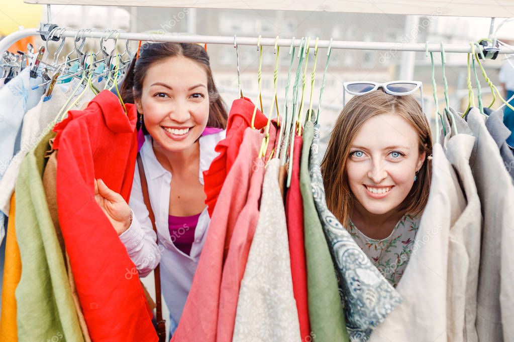 Two Smiling young women friends shopping for summer clothes together at outdoor flea market