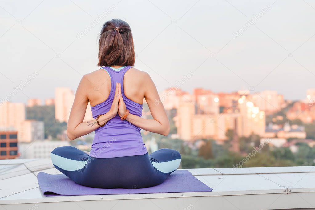 View of a healthy young sport woman practicing yoga sitting in lotos asana pose while meditating outdoors on the roof top against blue sky