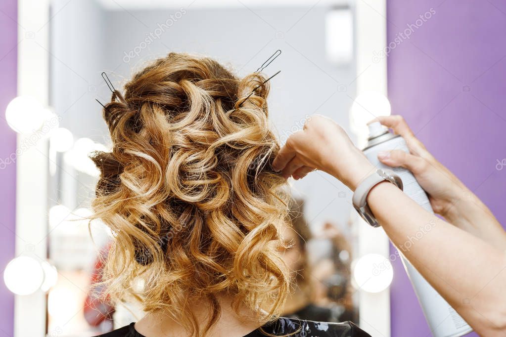 Female hairdresser makes hairstyle for young woman in beauty salon at the stylist workplace near the mirror