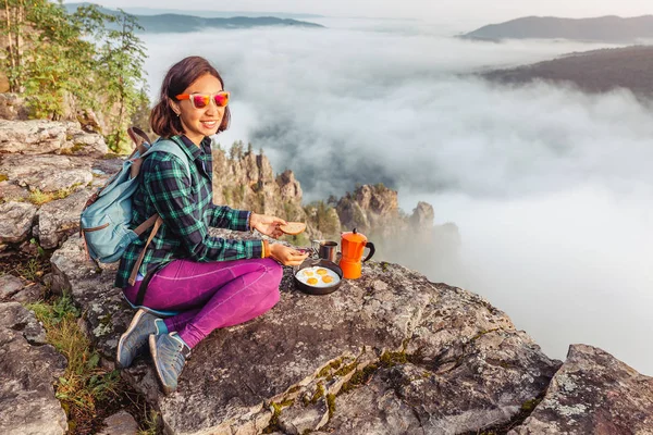 A woman traveler cooking and eating breakfast scrambled eggs and coffee at dawn in the misty mountains. Camping equipment and adventure concept